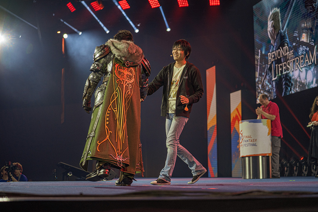 Final Fantasy 14 Producer and Director Naoki Yoshida dressed in a bastion costume shakes hands with Final Fantasy 7 Remake and Rebirth director Naoki Hamaguchi while on stage.