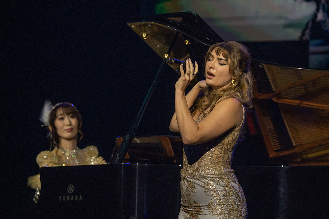 Singer Amanda Achen performs at the Piano Concert at Fan Festival 2026 in Berlin. She is wearing a gold and white sparkling form-fitting dress. Pianist Keiko performs accompanying music in the background.