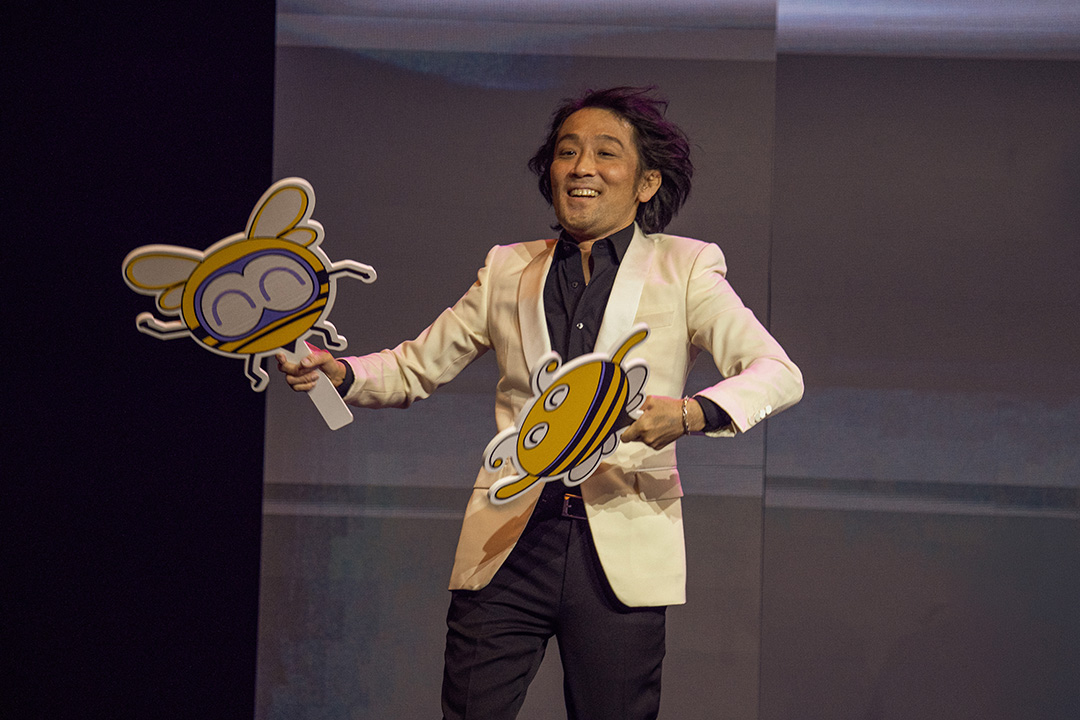 Final Fantasy 14 Sound Director Masayoshi Soken joyfully dancing while holding bee caricatures from Honey B Lovely's boss fight in-game. He is dressed in a white suit jacket and black suit trousers.