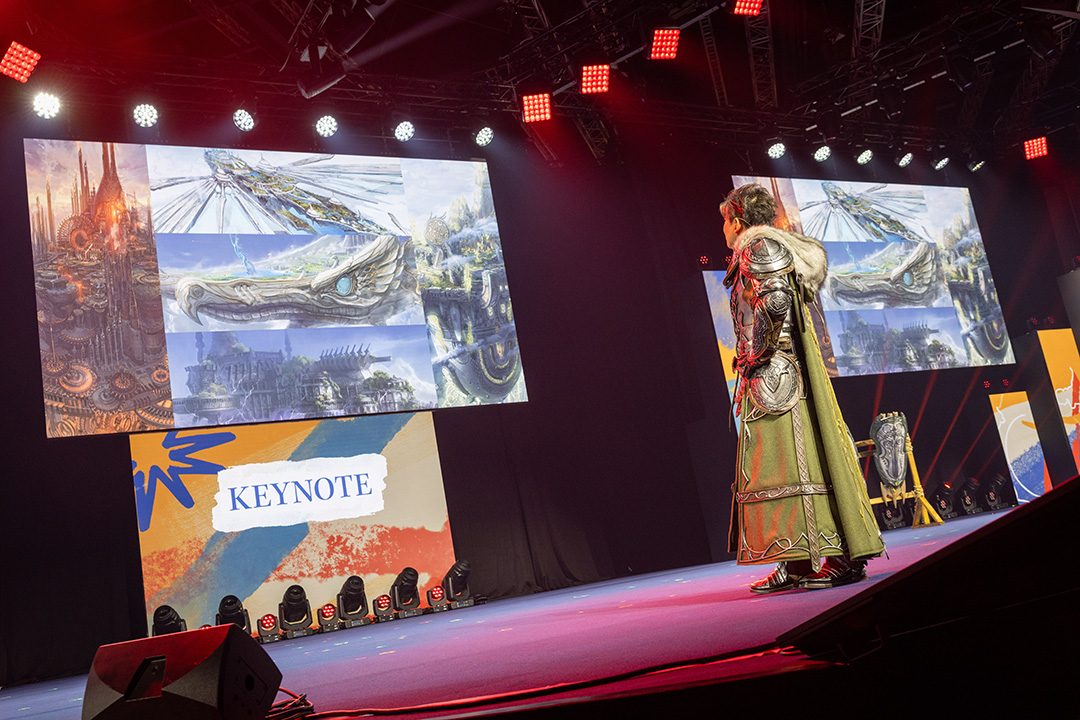 A photograph of Final Fantasy 14 Producer and Director Naoki Yoshida dressed in a bastion costume. He is on stage looking at concept art for Evercold during the keynote presentation.