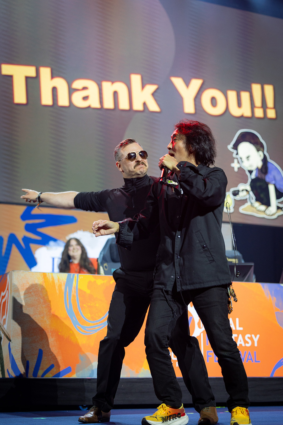 Final Fantasy 14 Composer and Sound Director Masayoshi Soken is being directed off stage by a Square Enix staff member impersonating a security guard in a joking manner.