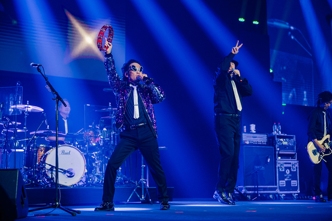 Masayoshi Soken and Koji Fox performing on stage at Fan Festival 2026 in Berlin. Soken is shaking a red tambourine in his right hand. 